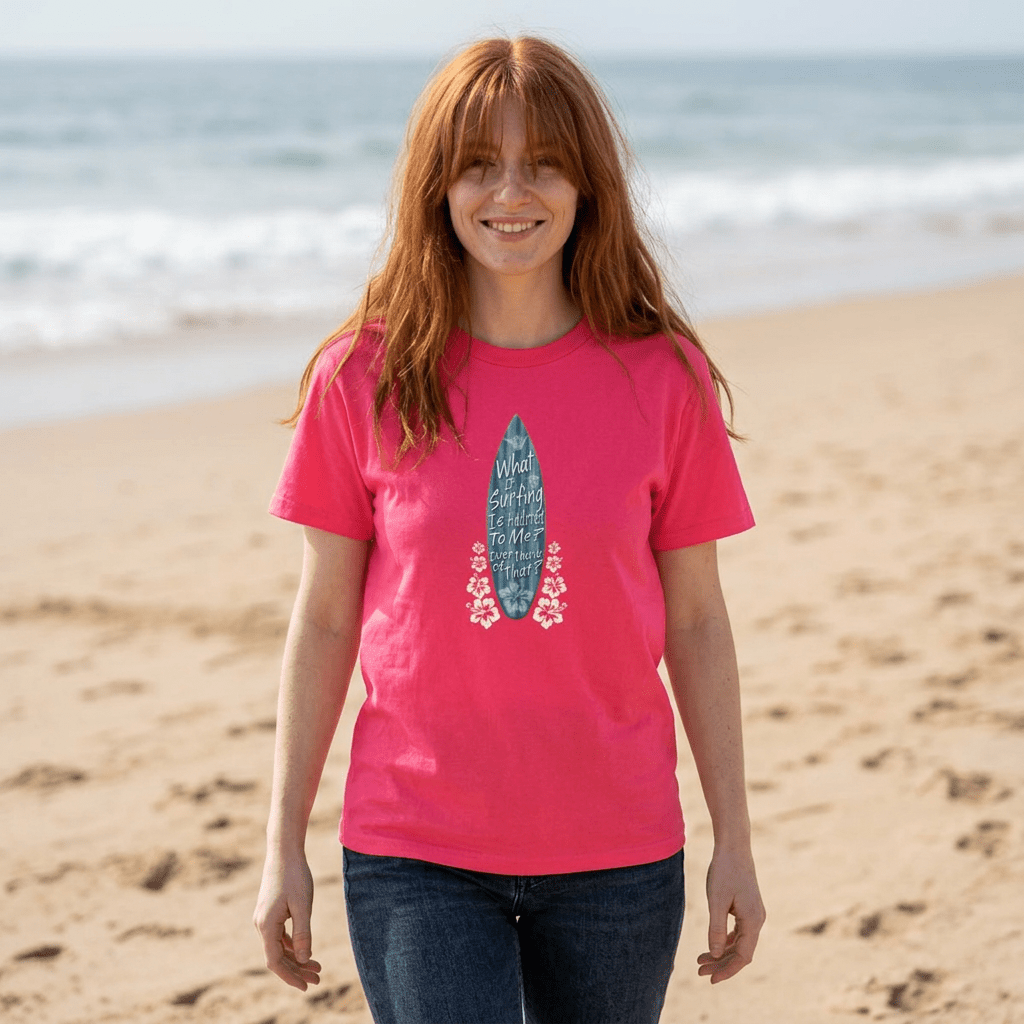 A woman with red hair stands on a sandy beach wearing a bright pink Shaka Wear t-shirt with a blue and pink surfboard graphic, paired with dark blue jeans.