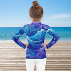 Little girl wearing a long-sleeve shirt with a blue and purple wave pattern on a beachside boardwalk