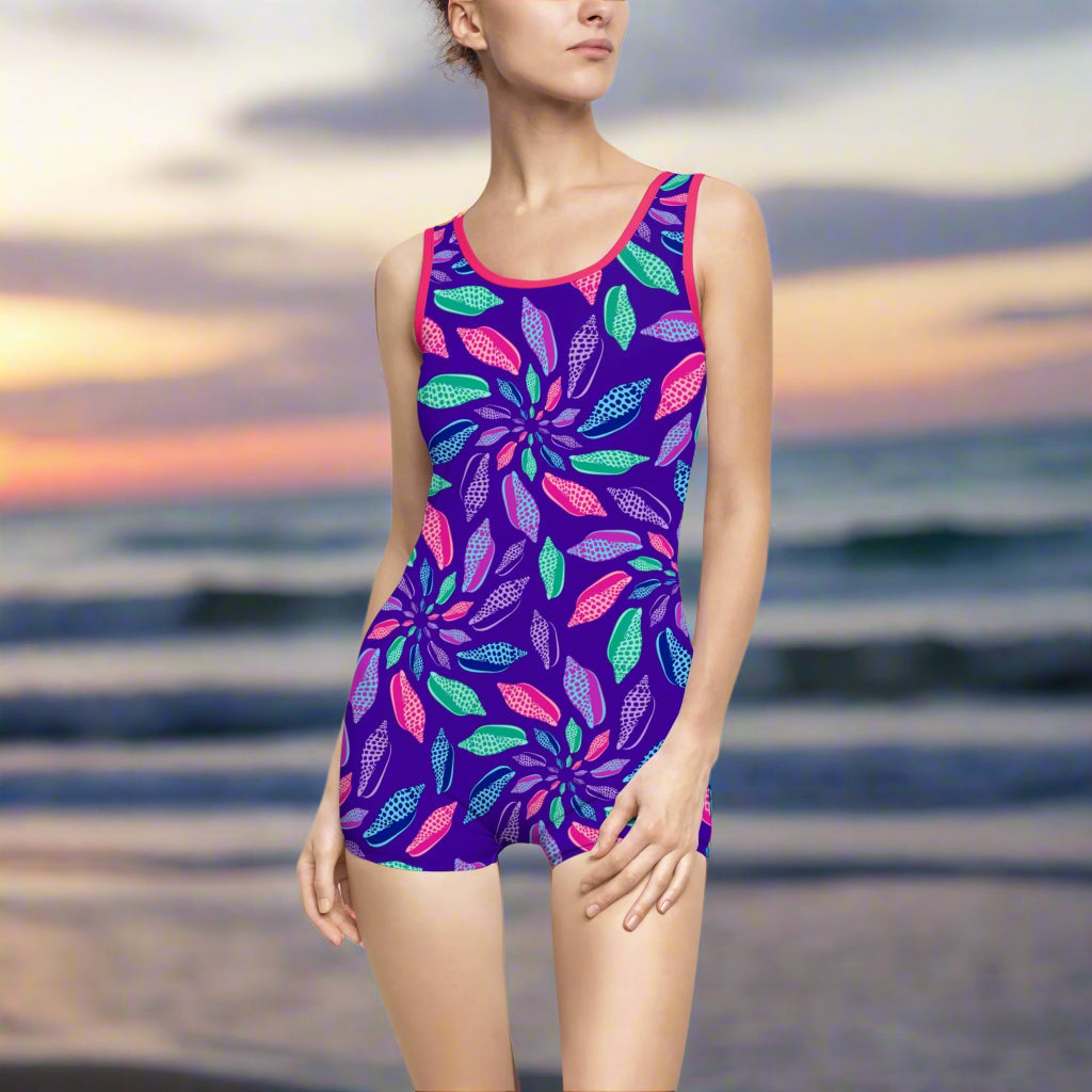 Model wearing blue one piece with pattern of junonia seashells and pink trim standing on a beach looking for shells