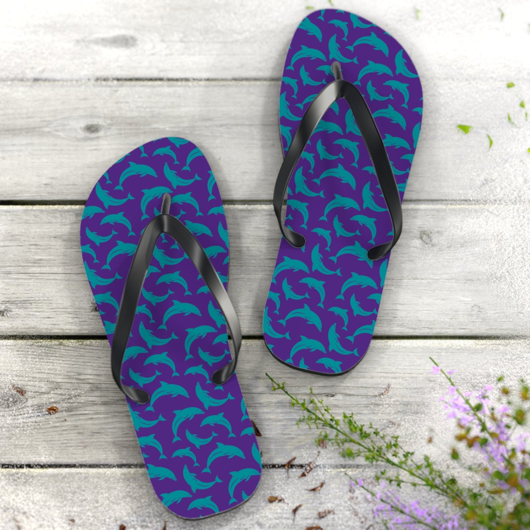Purple flip-flops with teal dolphin pattern on a wooden surface with flowers.