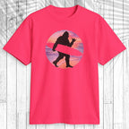 A bright pink short-sleeve t-shirt with a retro graphic of a Bigfoot silhouette carrying a pink surfboard inside a striped sunset circle, displayed on a white wood plank background.