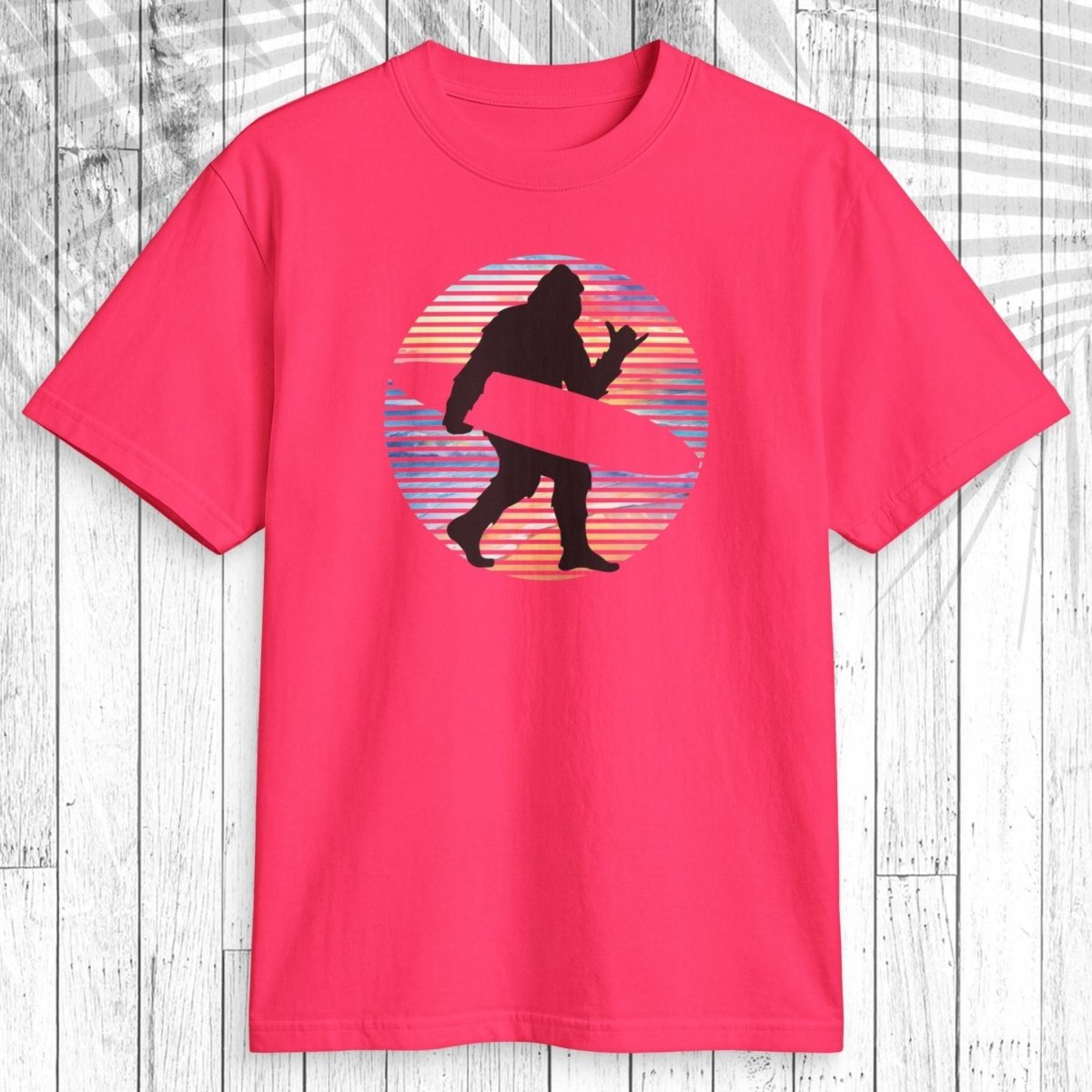 A bright pink short-sleeve t-shirt with a retro graphic of a Bigfoot silhouette carrying a pink surfboard inside a striped sunset circle, displayed on a white wood plank background.