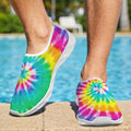 A Dude wearing slip-on water shoes with a bright rainbow tie-dye spiral pattern, standing on a pool deck with a swimming pool and outdoor background visible.