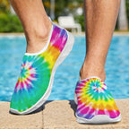 A Dude wearing slip-on water shoes with a bright rainbow tie-dye spiral pattern, standing on a pool deck with a swimming pool and outdoor background visible.