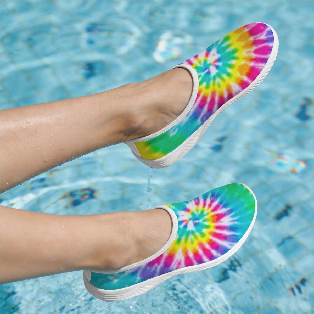 A pair of slip-on Tie Dye Beach Slippers with a vibrant rainbow tie-dye spiral pattern, shown being worn on feet dipping into a swimming pool.