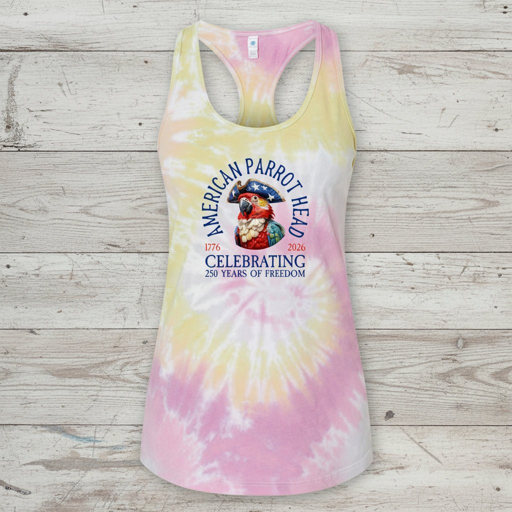 A racerback tank top with a pastel tie-dye pattern in pink, yellow, and white, featuring a graphic of an American parrot and the text 'American Parrot Breed Celebrating 250 Years of Freedom' on the front, displayed against a rustic light wood plank background.