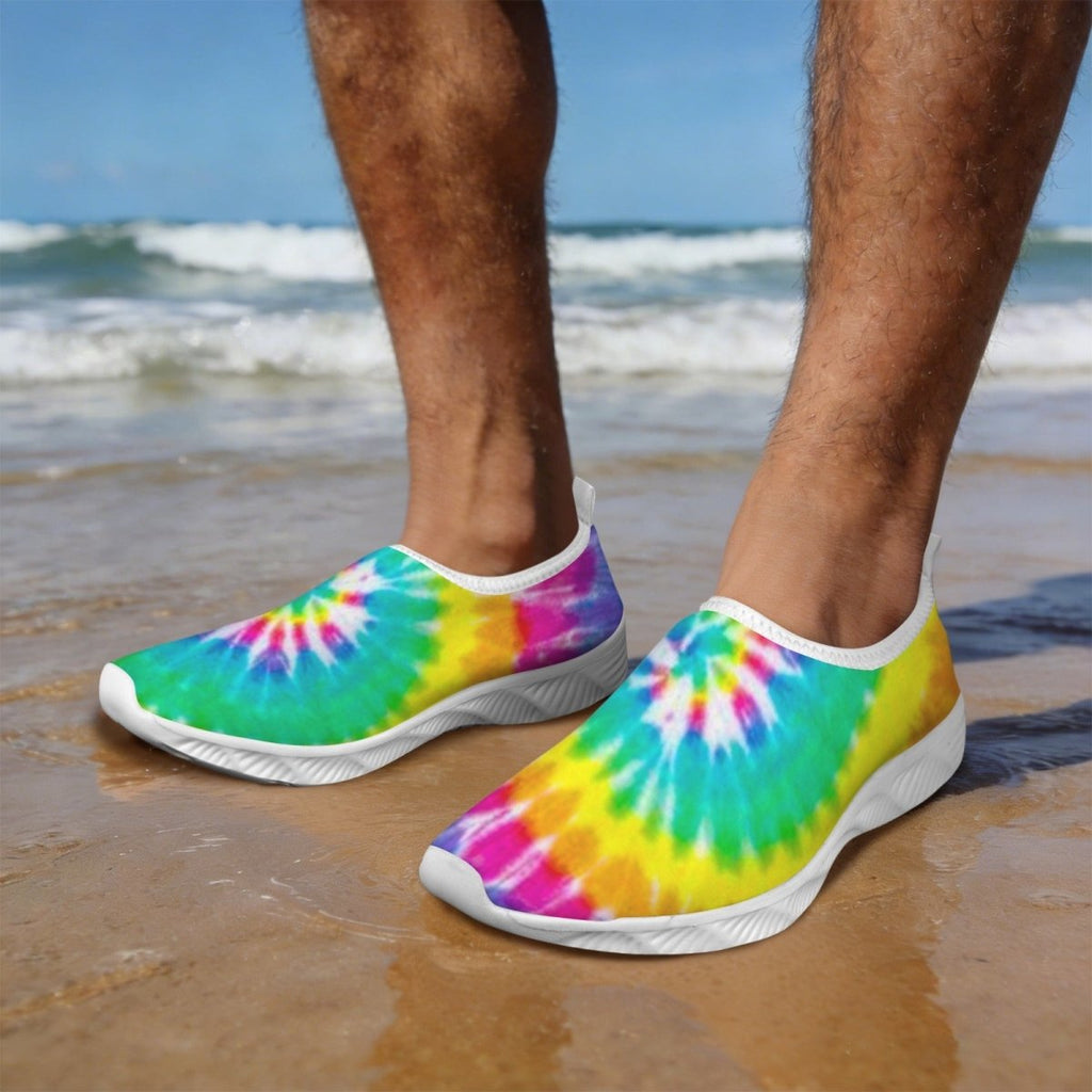 Dude standing on a sandy beach wearing Tie-Dye Water Slippers with a bright rainbow tie-dye pattern and white soles, with ocean waves and blue sky in the background.