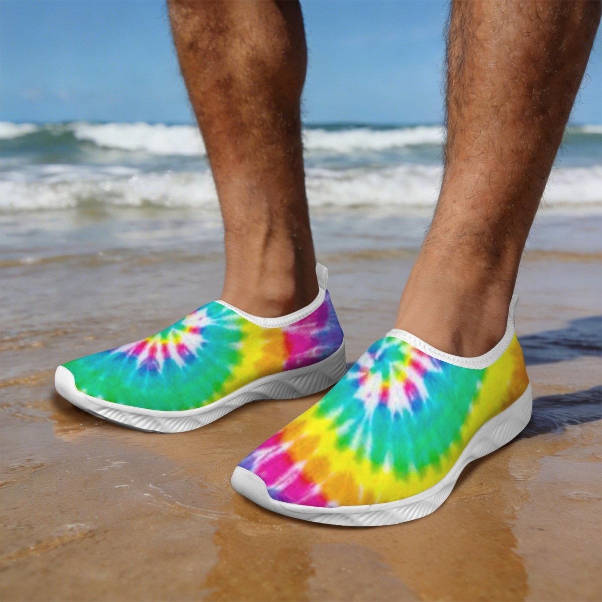 Dude standing on a sandy beach wearing Tie-Dye Water Slippers with a bright rainbow tie-dye pattern and white soles, with ocean waves and blue sky in the background.
