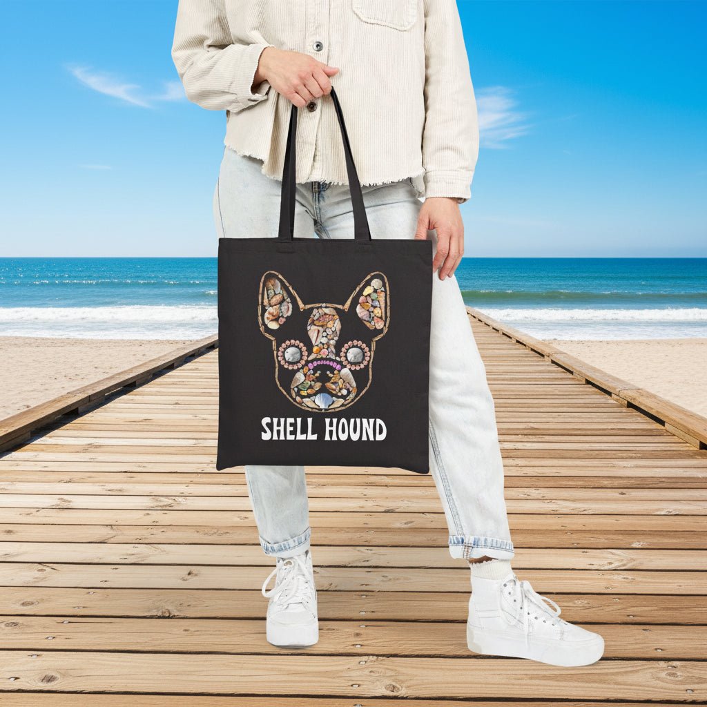 A person wearing a light beige jacket, light wash jeans, and white sneakers holds a black canvas tote bag featuring a colorful shell-art style dog illustration and the text 'SHELL HOUND'.