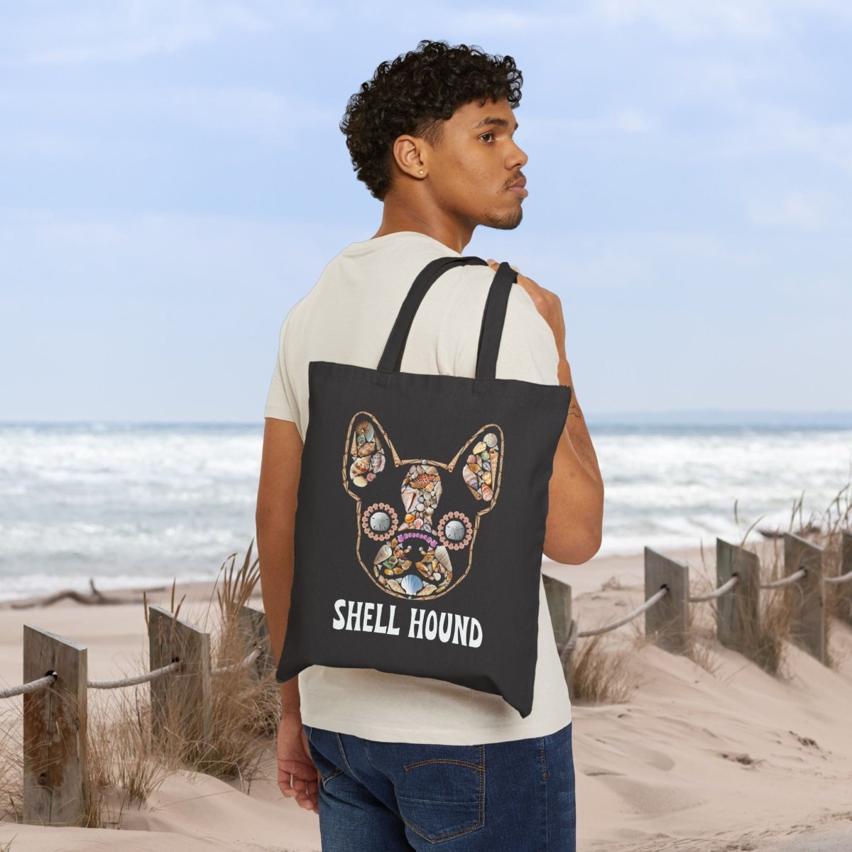 A man wearing a cream t-shirt and blue jeans stands on a sandy beach dune, carrying a black canvas tote bag over his shoulder. The bag features a colorful, detailed graphic of a French Bulldog's head with the text 'SHELL HOUND' printed below in white.