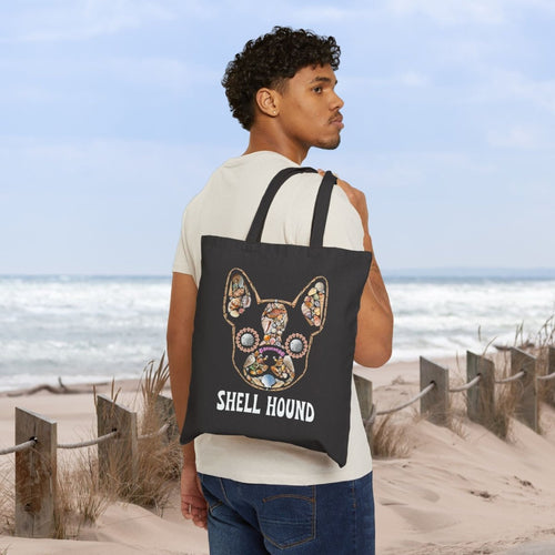 A man wearing a cream t-shirt and blue jeans stands on a sandy beach dune, carrying a black canvas tote bag over his shoulder. The bag features a colorful, detailed graphic of a French Bulldog's head with the text 'SHELL HOUND' printed below in white.