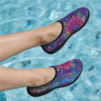 A pair of slip-on water shoes with a vibrant multicolor tribal pattern in purple, blue, pink, and orange, shown on a person's feet in a swimming pool.