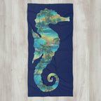 A rectangular beach towel with a dark navy blue background, featuring a large silhouette of a seahorse filled with a tropical palm tree and wave pattern in shades of teal, green, and yellow.