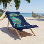 A navy blue beach towel with a colorful tropical map of Madagascar print, draped over a wooden lounge chair on a sandy beach with palm trees and the ocean in the background.