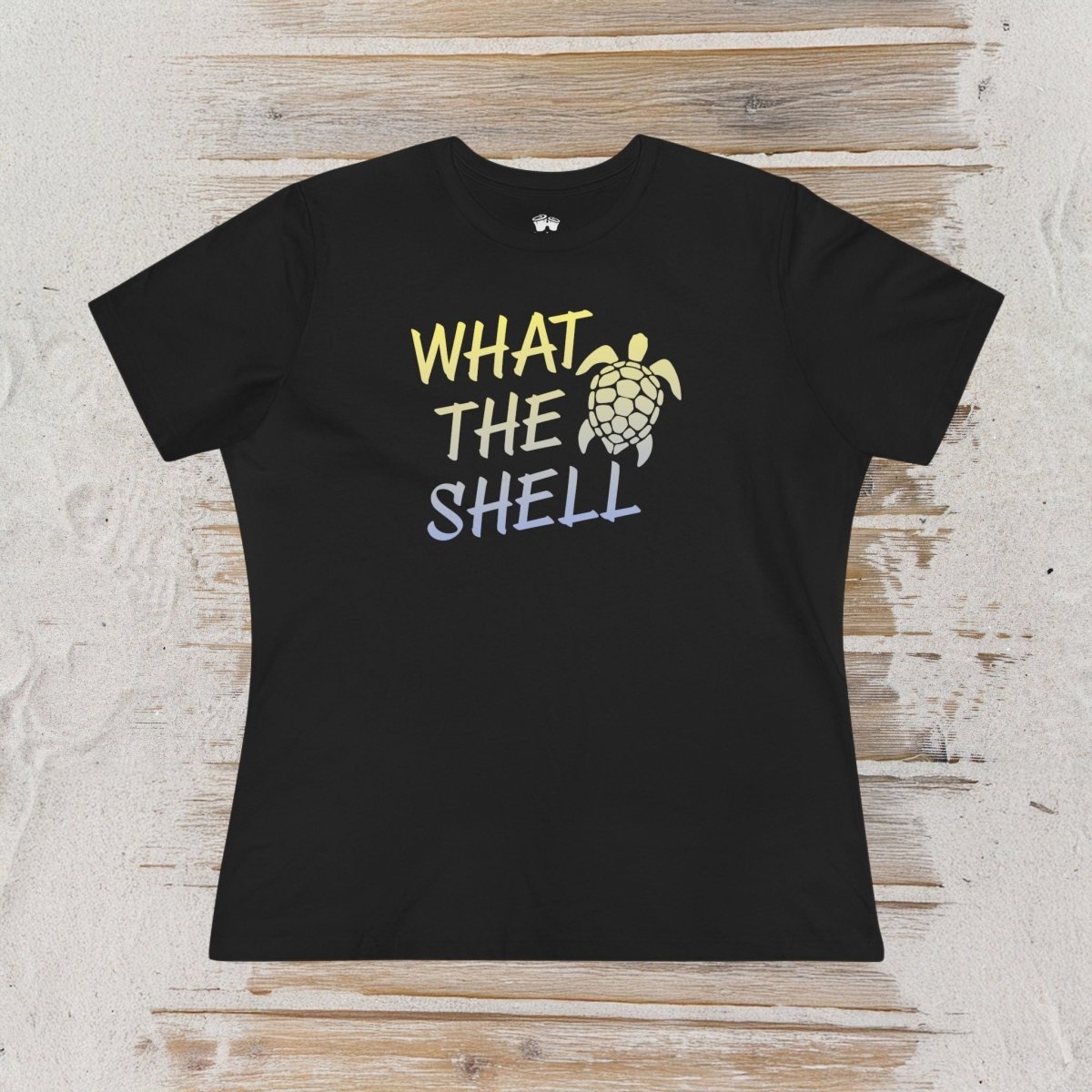 A black short-sleeve t-shirt with a graphic print reading 'WHAT THE SHELL' in yellow, white, and blue lettering, accompanied by a sea turtle illustration. The shirt is laid flat on a light-colored, rustic wood plank background.