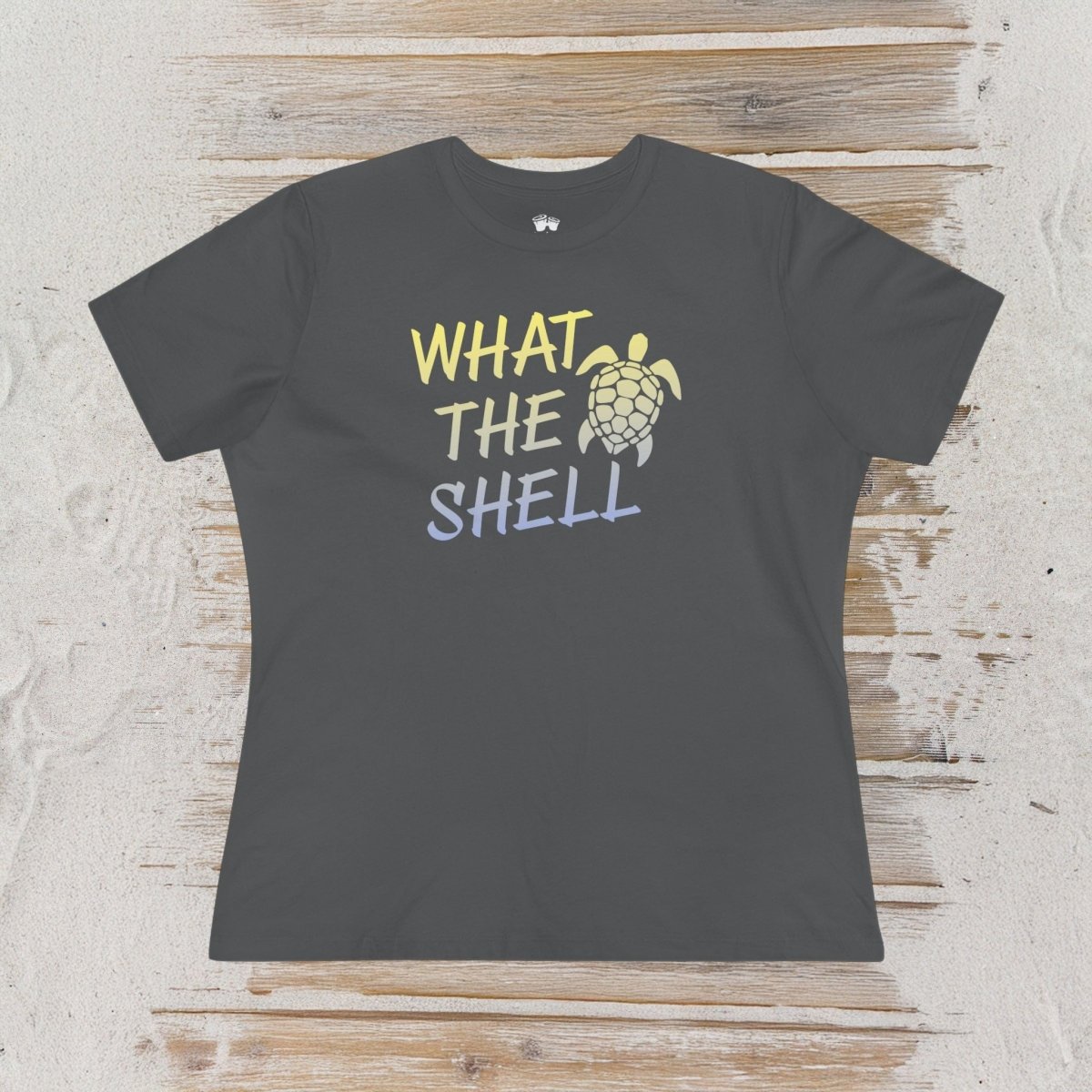 A dark charcoal gray short-sleeve t-shirt with a graphic print reading 'WHAT THE SHELL' in yellow, white, and light blue, alongside a sea turtle illustration. The shirt is laid flat on a rustic light wood background.