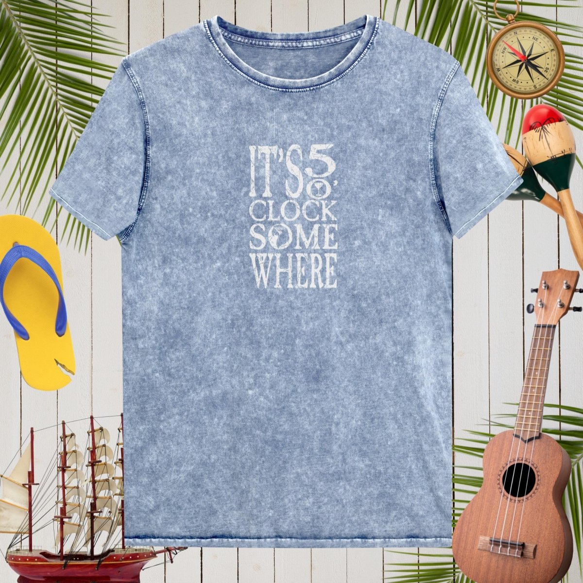 Blue t-shirt with 'It's 5 o'clock somewhere' text on a wooden background with tropical elements.