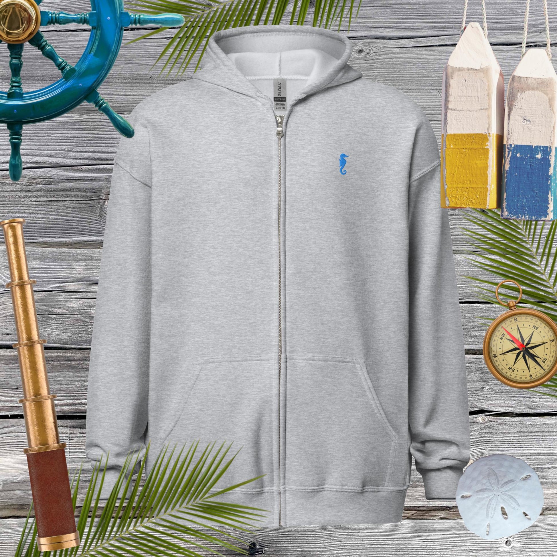 Unisex heavy blend zip hoodie with seahorse embroidery, set against nautical decor and palm leaves for a cozy vibe.