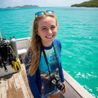 A smiling young woman stands on a boat deck wearing a blue long-sleeve rash guard with a colorful ocean scene print, sunglasses on her head, and a camera around her neck, with turquoise Virgin Island water and distant islands in the background.