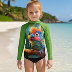 A young girl with a bun stands on a tropical beach wearing a long-sleeve swim rash guard. The shirt features bright green sleeves and a colorful print of a rainbow-colored parrot on the front, paired with black swim bottoms.