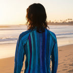 A teen surfer dude standing on Venice Beach at sunset, viewed from behind, wearing a long-sleeve striped shirt in shades of blue, teal, and purple.