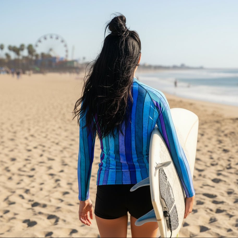 A teenage surfer girl on Venice Beach, seen from behind, wearing a blue and purple vertically striped long-sleeve rash guard and carrying a white surfboard.