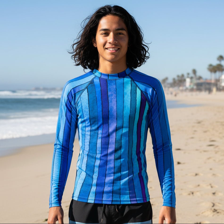 A teenage dude standing on Venice Beach wearing a long-sleeve blue striped rash guard and black swim trunks.