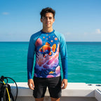A young teen stands on a boat ,off the coast of Vero Beach, wearing a long-sleeve blue Nemo's Brother swim rash guard with a colorful clownfish and coral print, paired with black swim shorts against a turquoise ocean background.