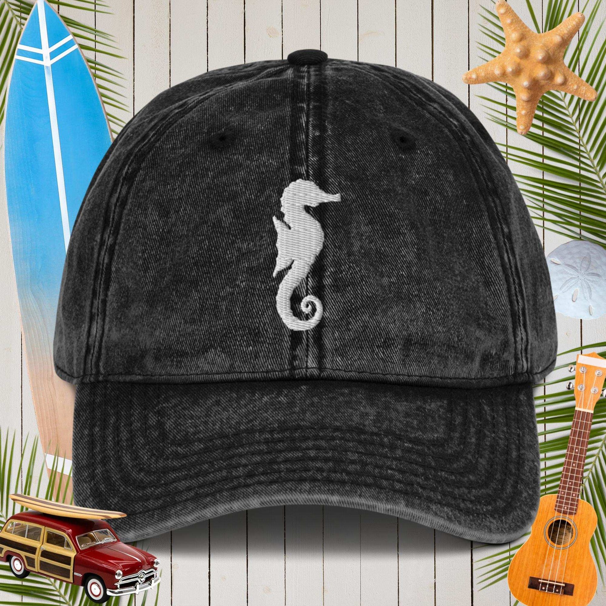 Black cap with a white seahorse design on a wooden background with beach-themed items.