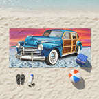 A rectangular beach towel featuring a vintage blue wood-paneled station wagon on a sandy beach under a colorful sunset sky.