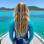 A tween girl with long, wavy blonde hair stands on a boat deck facing turquoise sea in the Virgin Islands water and distant green hills, wearing a blue long-sleeve top with a dark blue and black abstract print on the lower portion.