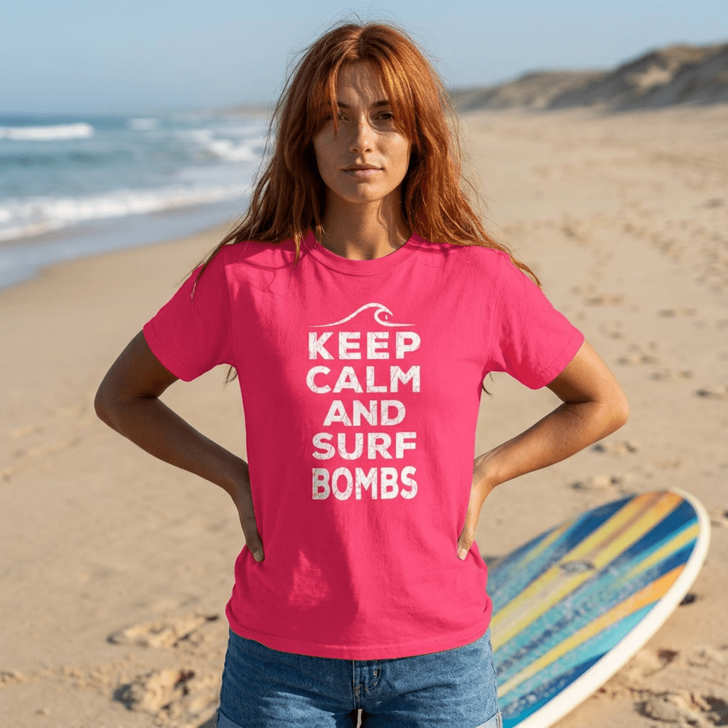A woman with red hair stands on a sandy beach wearing a bright pink t-shirt with the white text 'KEEP CALM AND SURF BOMBS' and blue denim shorts. A colorful surfboard is on the sand next to her, with the ocean and dunes in the background.
