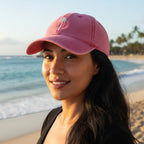 A woman wearing a pink baseball cap with a small white palm tree logo, photographed outdoors on a beach with ocean waves and sand in the background.
