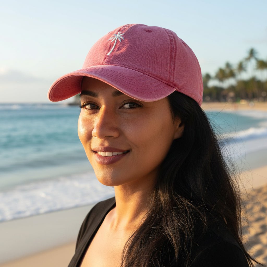 A woman wearing a pink baseball cap with a small white palm tree logo, photographed outdoors on a beach with ocean waves and sand in the background.