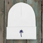 A white ribbed knit beanie hat with a folded cuff, featuring a small navy blue embroidered palm tree on the front, set against a rustic wood background.