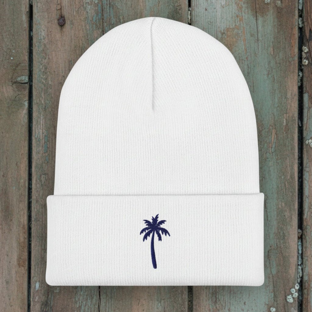 A white ribbed knit beanie hat with a folded cuff, featuring a small navy blue embroidered palm tree on the front, set against a rustic wood background.