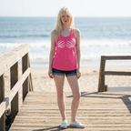 A woman stands on a wooden boardwalk at the beach wearing a bright pink tank top with a white skeleton hand graphic, dark denim shorts, and white sneakers.