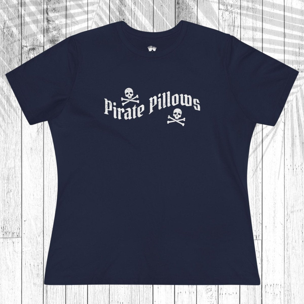 A navy blue short-sleeve t-shirt with a white graphic reading 'Pirate Pillows' and skull-and-crossbones icons, laid flat on a white wood plank background.