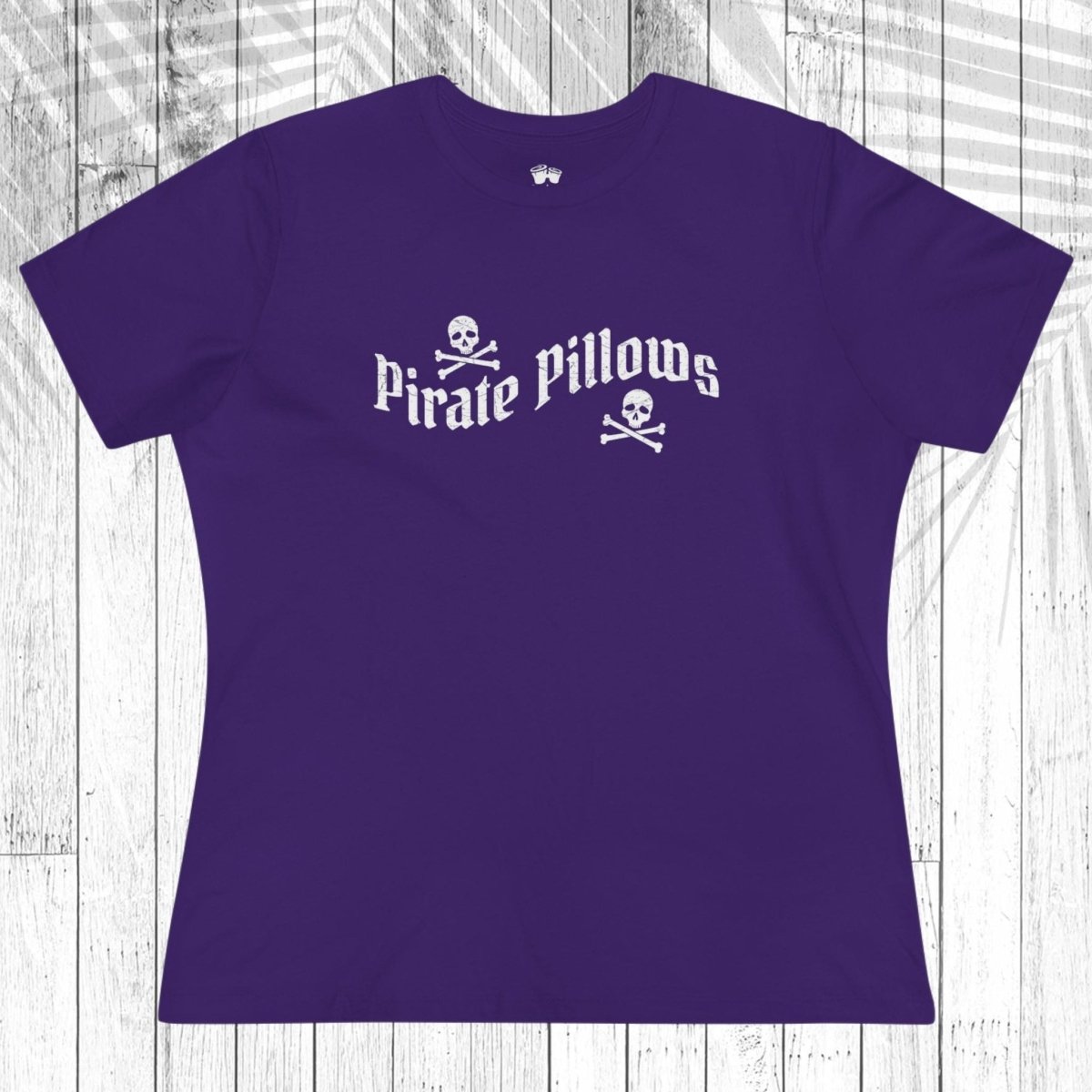 A navy blue short-sleeve t-shirt with a white graphic reading 'Pirate Pillows' and skull-and-crossbones icons, laid flat on a white wood plank background.