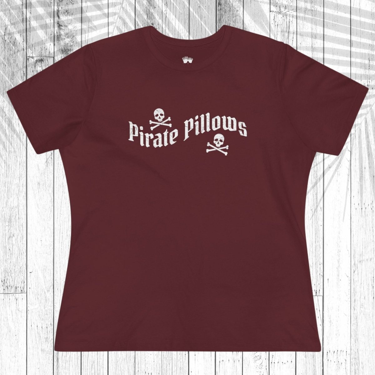A cardinal blue short-sleeve t-shirt with a white graphic reading 'Pirate Pillows' and skull-and-crossbones icons, laid flat on a white wood plank background.