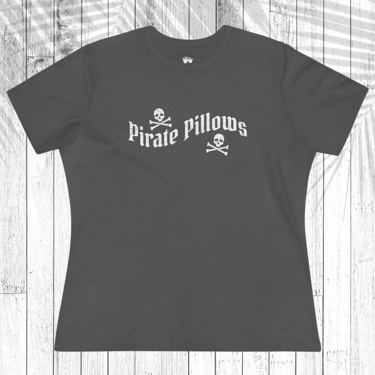 A green short-sleeve t-shirt with a white graphic reading 'Pirate Pillows' and skull-and-crossbones icons, laid flat on a white wood plank background.