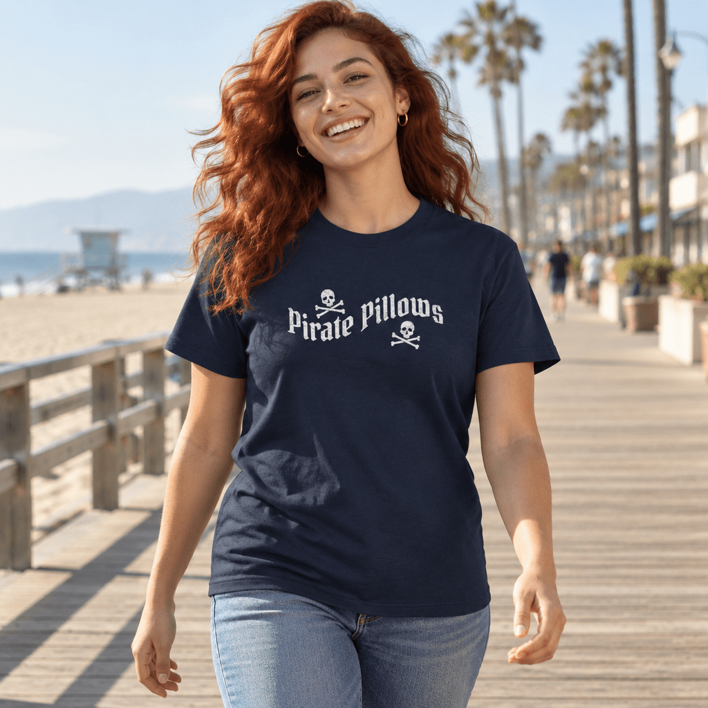 A woman with long red hair walking on a sunny wooden boardwalk at the beach, wearing a navy blue t-shirt with a white 'Pirate Pillows' graphic and light blue jeans.