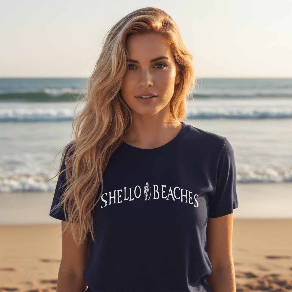 A navy blue short-sleeve crewneck t-shirt with 'SHELLO BEACHES' printed in white arched text across the chest, laid flat on a light wood plank background.