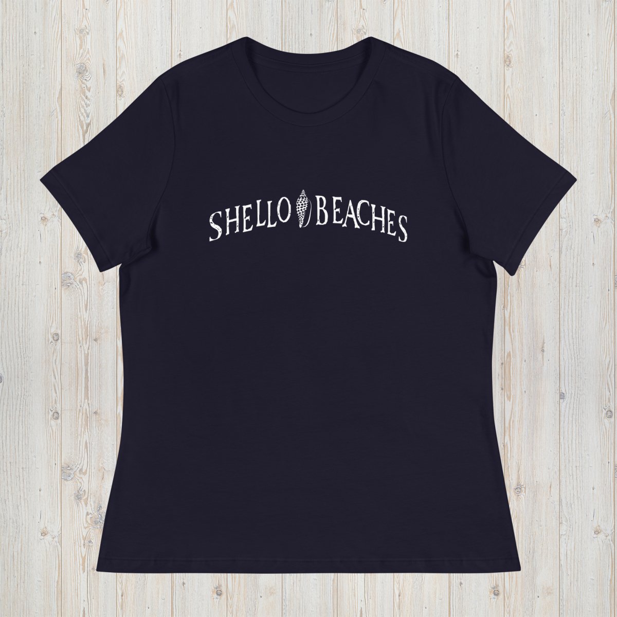 A navy blue short-sleeve crewneck t-shirt with 'SHELLO BEACHES' printed in white arched text across the chest, laid flat on a light wood plank background.