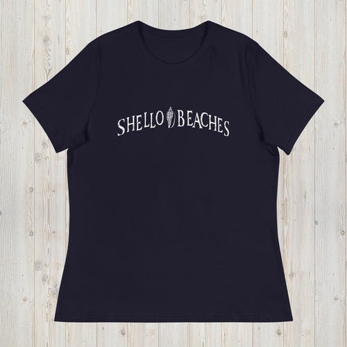 A navy blue short-sleeve crewneck t-shirt with 'SHELLO BEACHES' printed in white arched text across the chest, laid flat on a light wood plank background.
