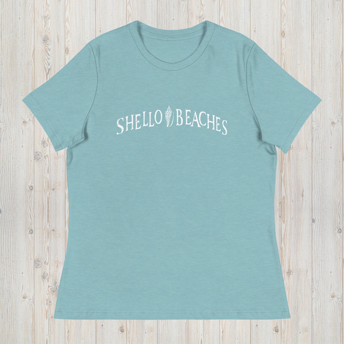 A heathered teal short-sleeve t-shirt with 'SHELLO BEACHES' printed in white across the chest, laid flat on a light wood plank background.