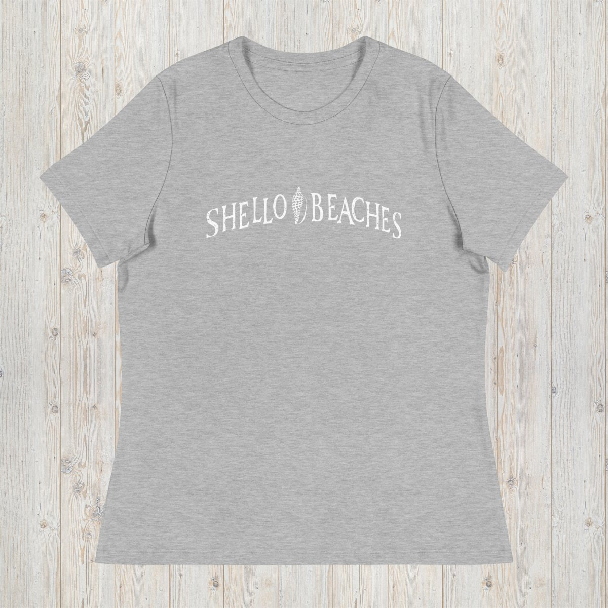 A heather gray short-sleeve t-shirt with 'SHELLO BEACHES' printed in white across the chest, laid flat on a light wood plank background.