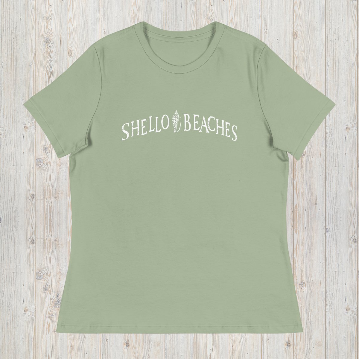A sage green short-sleeve t-shirt with 'SHELLO BEACHES' printed in white across the chest, laid flat on a light wood plank background.