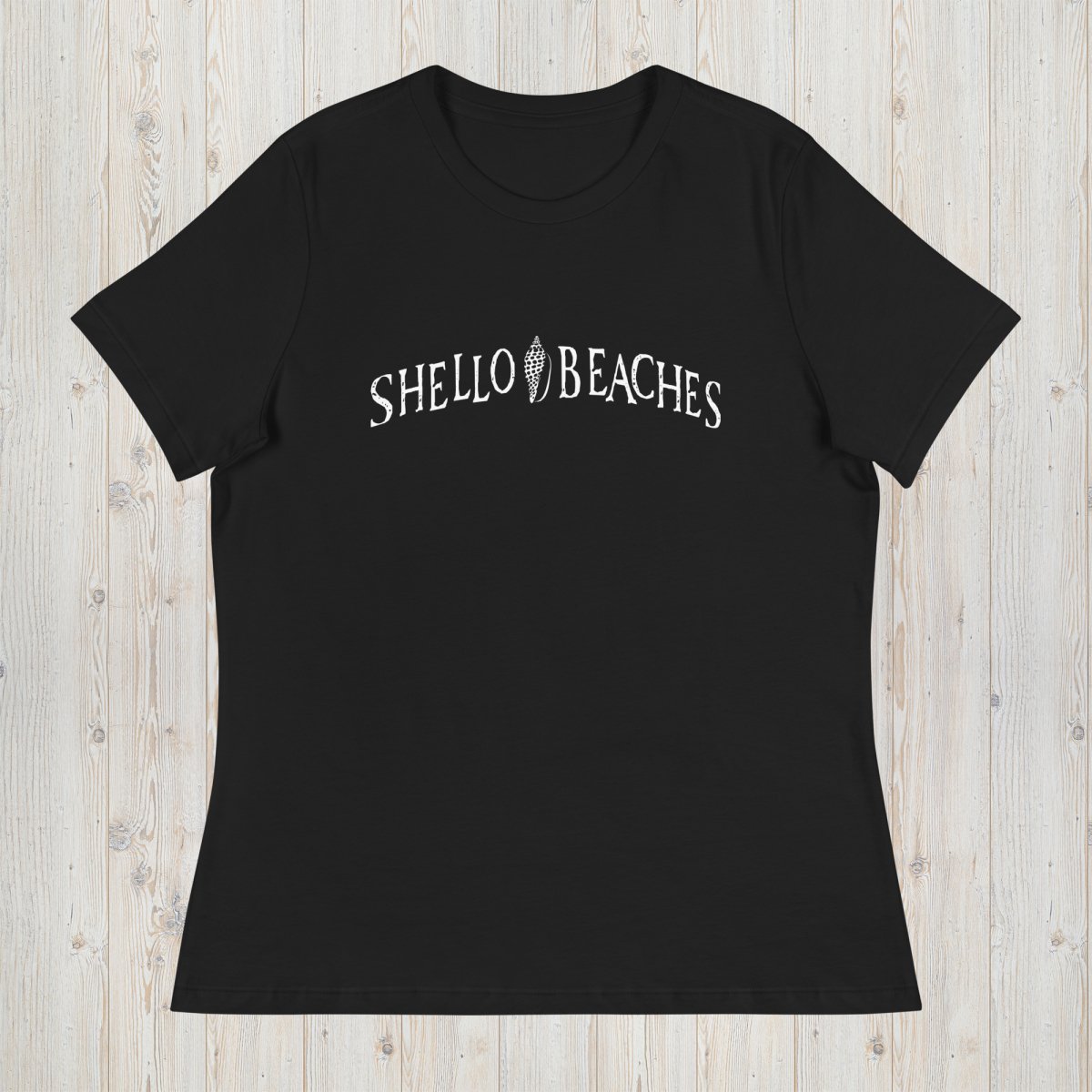 A black short-sleeve crewneck t-shirt with 'SHELLO BEACHES' printed in white arched text across the chest, laid flat on a light wood plank background.